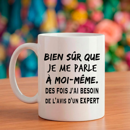 Mug blanc Bruhle avec texte noir : Bien sûr que je me parle à moi-même, des fois j'ai besoin de l'avis d'un expert, entouré d'un décor de café chaleureux.