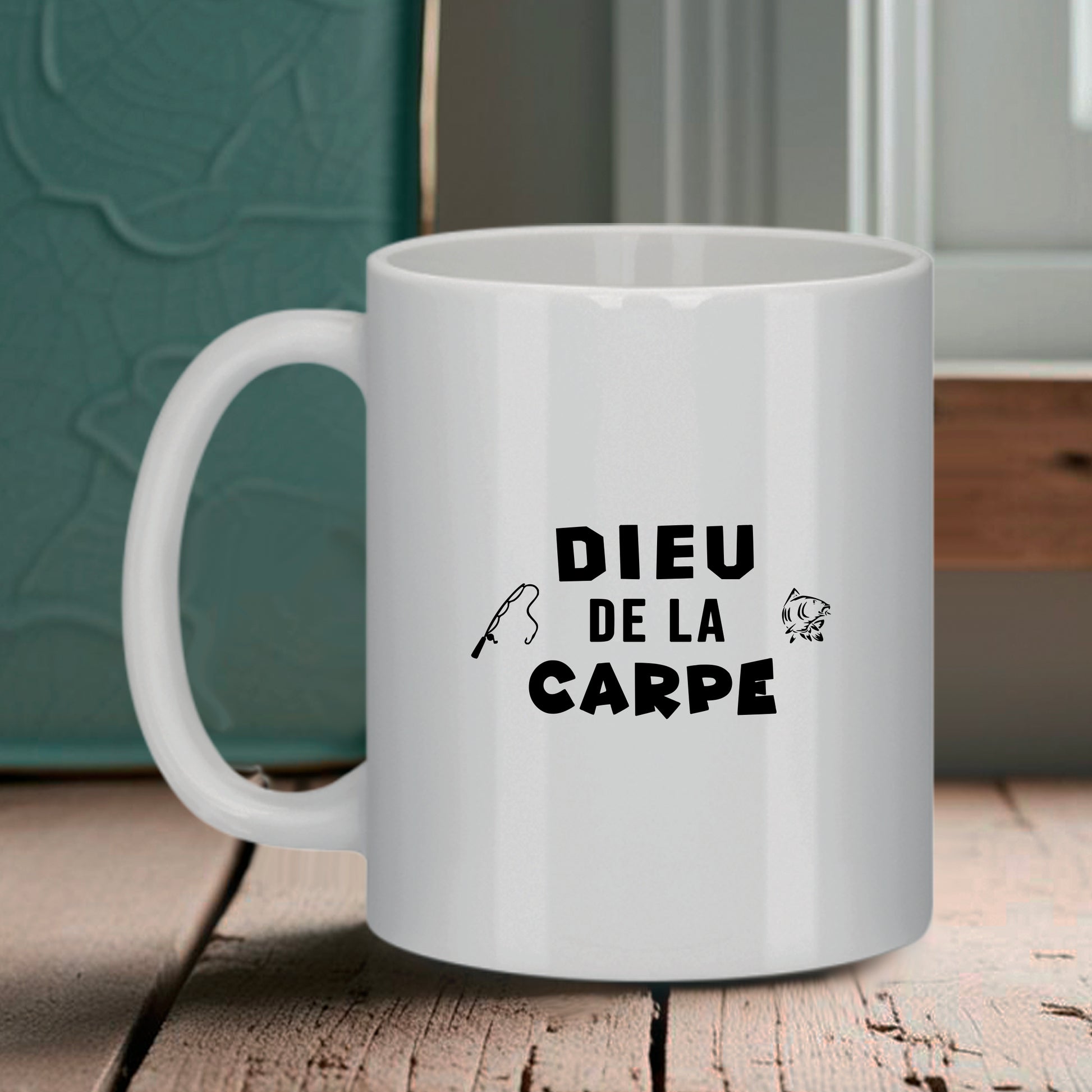 Mug blanc Bruhle avec texte noir "DIEU DE LA CARPE" entouré d'une petite canne à pêche et d'un poisson, posé sur une table en bois.