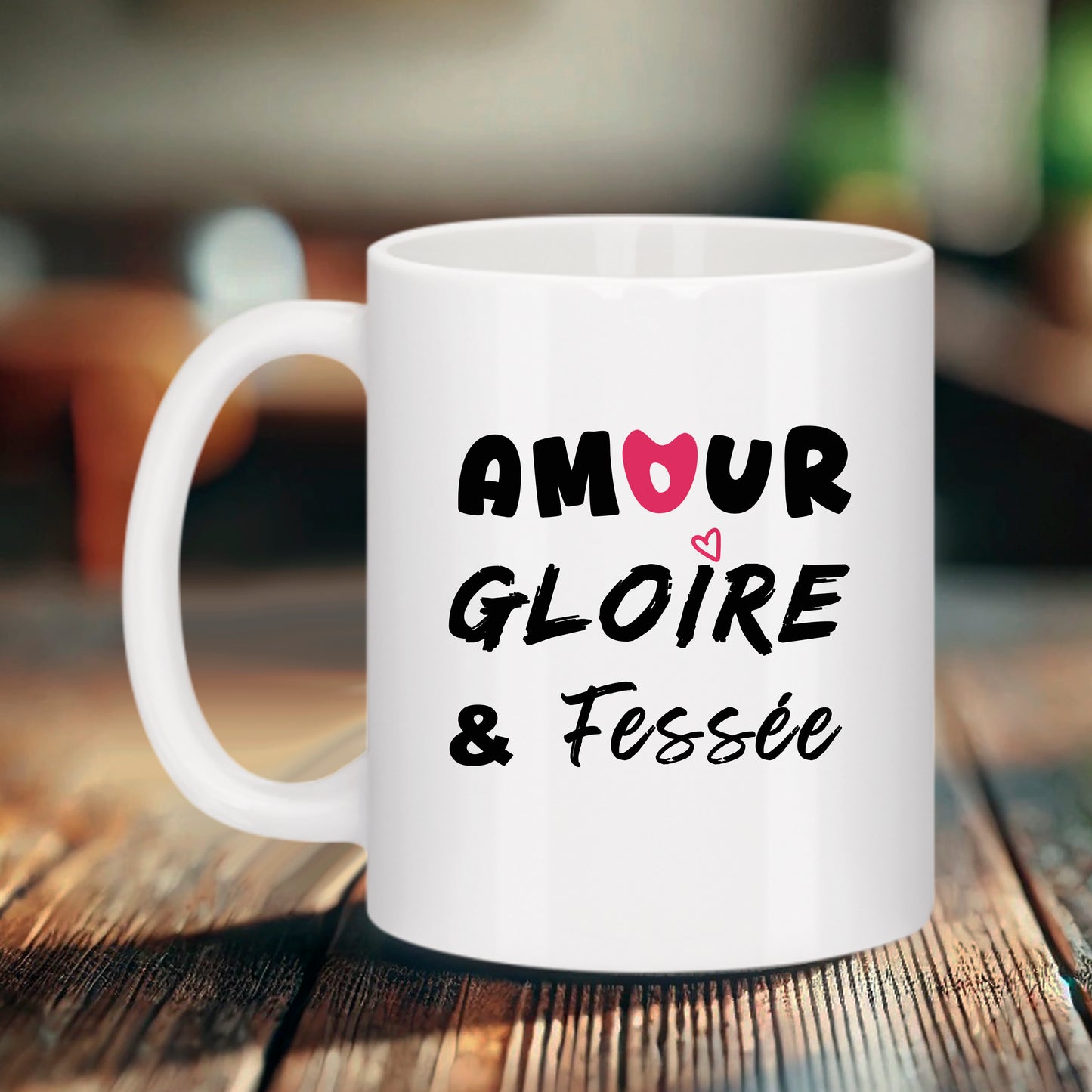 Mug blanc Bruhle présenté sur un fond boisé, affichant le texte "AMOUR" avec un cœur rose, "GLOIRE" et "& Fessée" dans une police manuscrite élégante, décoré d'un petit cœur rose filaire.