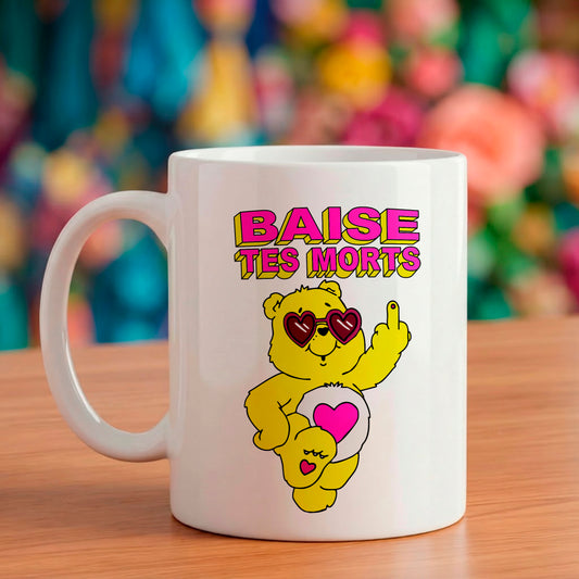 Mug en céramique blanc illustré d'un nounours jaune portant des lunettes en forme de cœur faisant un doigt d'honneur avec le texte 'BAISE TES MORTS' écrit en style rétro au-dessus.