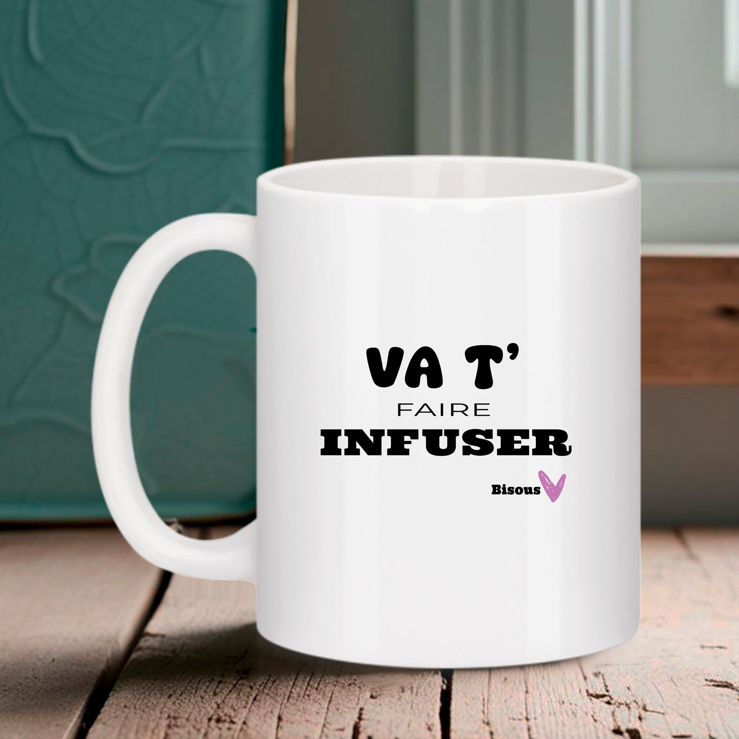 Mug blanc Bruhle avec texte noir "VA T' FAIRE INFUSER" et un petit "Bisous" accompagné d'un cœur rose, posé sur une table en bois.