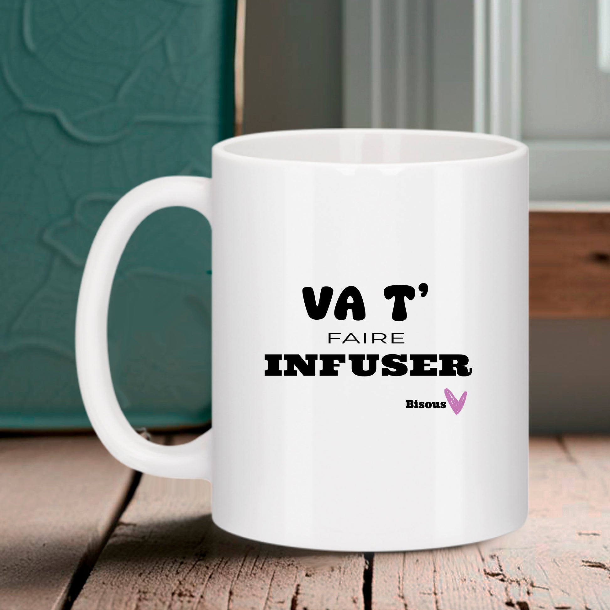 Mug blanc Bruhle avec texte noir "VA T' FAIRE INFUSER" et un petit "Bisous" accompagné d'un cœur rose, posé sur une table en bois.
